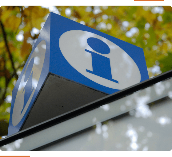 Blue information sign with a white background and an "i" symbol, mounted on a building against a backdrop of green and yellow foliage.
