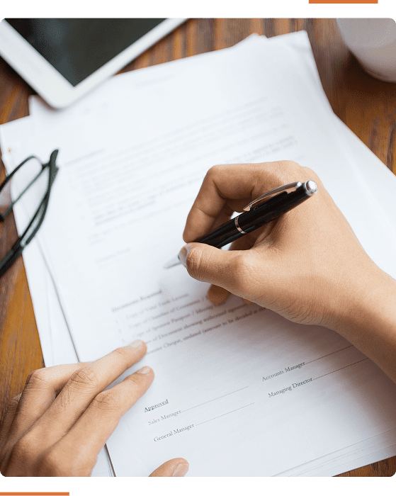 9 A person is signing papers on a wooden desk with a black pen. Glasses and a tablet are placed nearby.