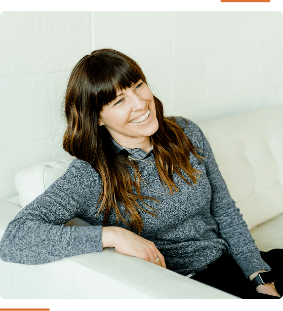 Person with long brown hair and bangs, wearing a gray sweater, sits on a white couch and smiles, looking to the side.