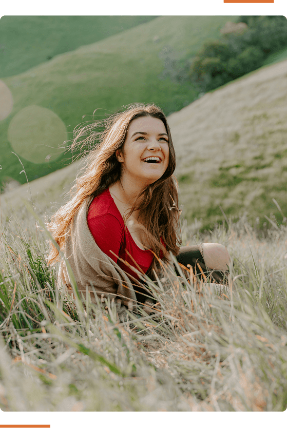 A person with long hair is sitting in a grassy field, smiling broadly, with hills and greenery in the background.