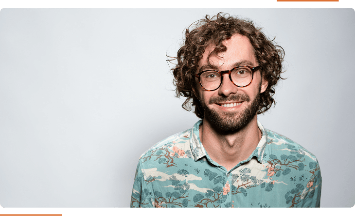 A man with curly hair and glasses smiles while wearing a floral-patterned shirt against a plain background.