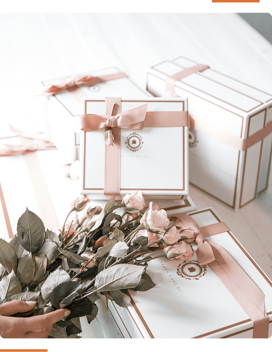 White gift boxes wrapped with pink ribbon are stacked together. A hand holds a bouquet of light pink roses in front of the boxes.