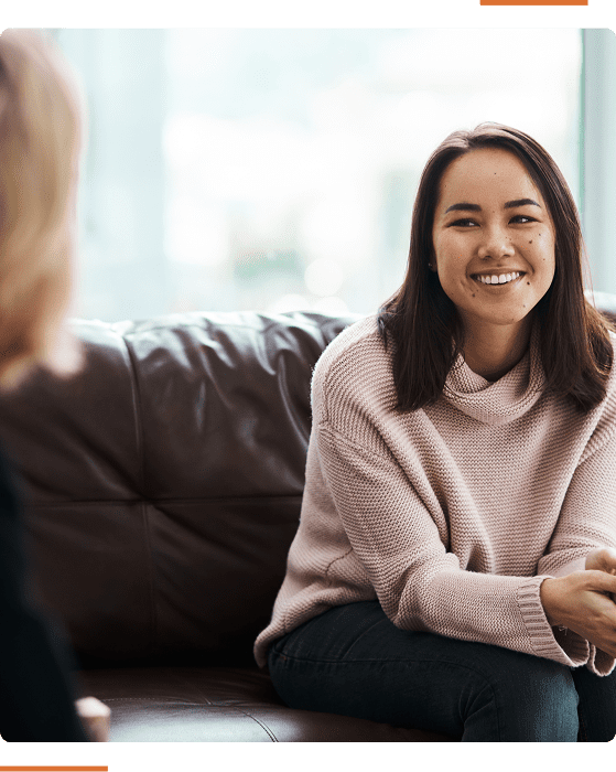 A woman in a light sweater and jeans sits on a brown sofa, smiling and talking to another person blurred in the foreground.
