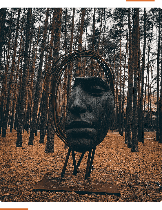 A large face sculpture stands amidst tall pine trees, surrounded by fallen leaves.