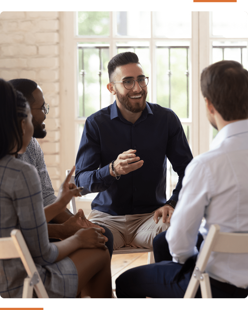 A group of four people seated in a circle indoors, having a discussion. One person is speaking while others listen attentively.