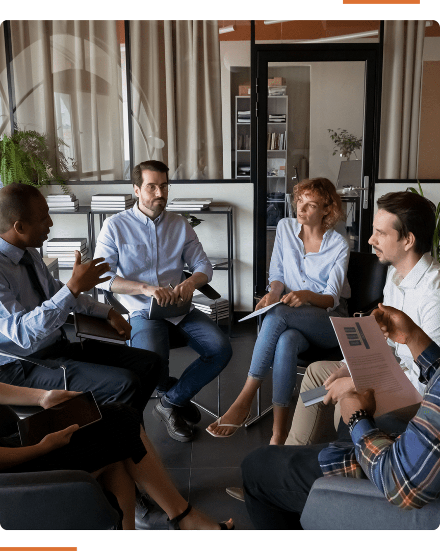 A group of people sitting in a circle in an office setting, engaged in a discussion, holding papers and digital tablets.