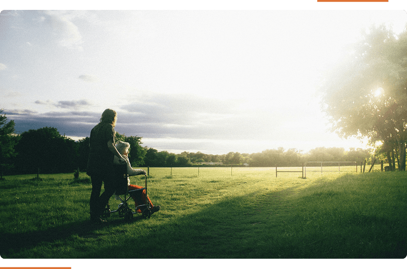 A person pushes another in a wheelchair along a grassy path at sunset, with trees and a fence in the background.