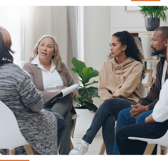A group of people sit in chairs having a discussion in a room with a plant and bookshelf in the background.