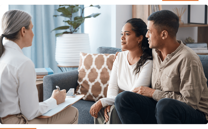A couple sits on a sofa, engaged in conversation with a professional holding a clipboard.