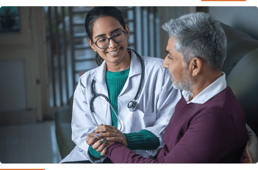 A healthcare professional in a white coat and stethoscope talks with an older person sitting on a couch. They are facing each other, and the professional is smiling.