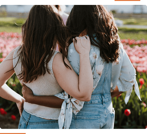 Two people stand with arms around each other, facing away, in a tulip field. Both are wearing casual clothing with floral and denim patterns.