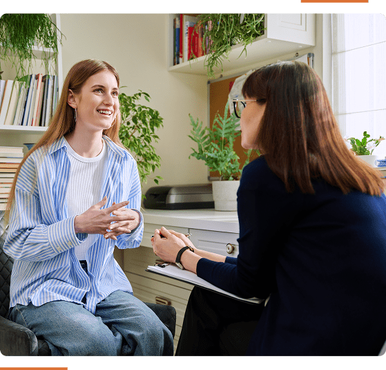 Two people having a conversation in a well-lit room with plants and shelves in the background. One is speaking, the other listening attentively.