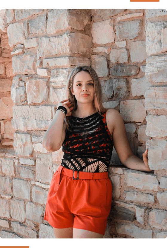 A person wearing a black and red top and orange shorts leans against a stone wall.