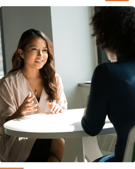 Two people sitting at a round table, engaged in conversation near a window. One person is speaking with expressive gestures.