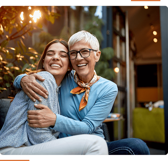Two people sit closely together. One person has short gray hair and glasses, the other has long dark hair. Both are smiling while hugging outside near plants.